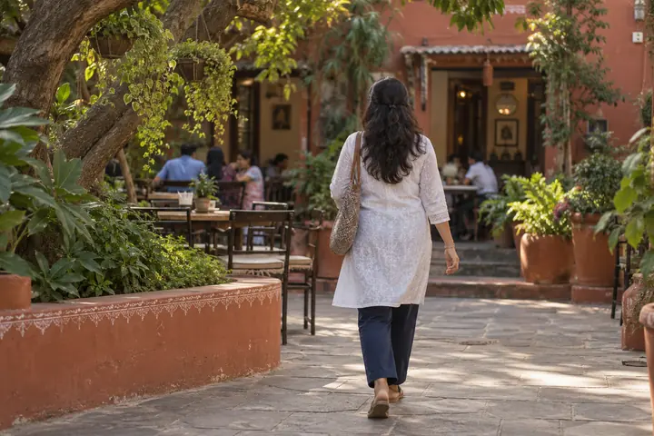 Fictional adult woman viewed from behind: walking through a public cafe courtyard with hanging plants and low terracotta walls; afternoon