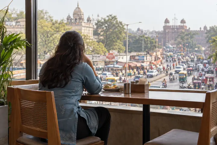 Fictional adult woman viewed from behind: seated at a public restaurant window overlooking a busy regional street; lunchtime