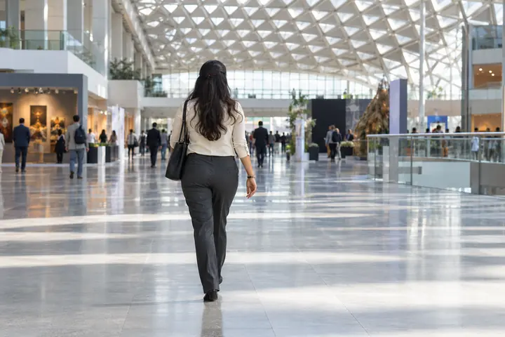 Fictional adult woman viewed from behind: walking across a spacious public exhibition concourse with geometric ceiling; midday