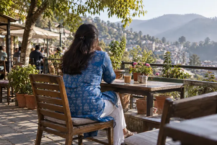 Fictional adult woman viewed from behind: seated from behind at a neighborhood tea shop terrace with potted plants; morning