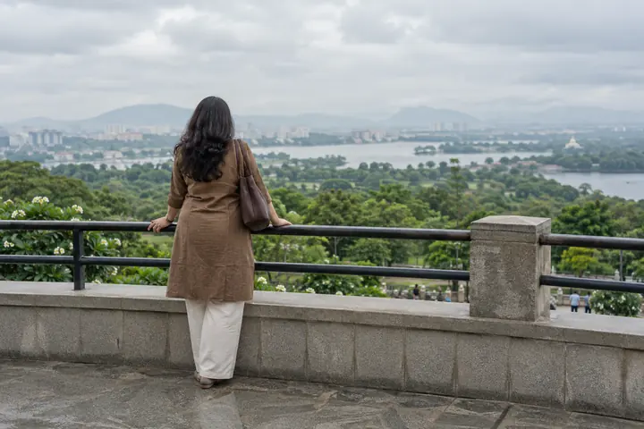 Fictional adult woman viewed from behind: looking across greenery from a public urban viewing terrace; overcast morning